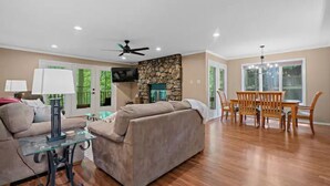 A living room with a stone fireplace, a dining area with wooden furniture, and a ceiling fan.