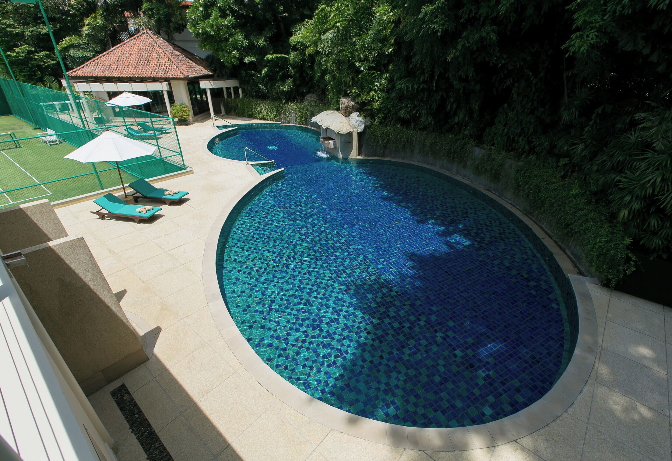 Una piscina con borde de azulejos, rodeada por una valla verde y sillas de descanso.