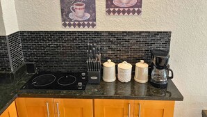 A kitchen with a black tiled backsplash, a black stovetop, and wooden cabinets. There are two coffee dispensers and a coffee maker on the counter. - Elegant Condo - European Charm (Palm Coast)
