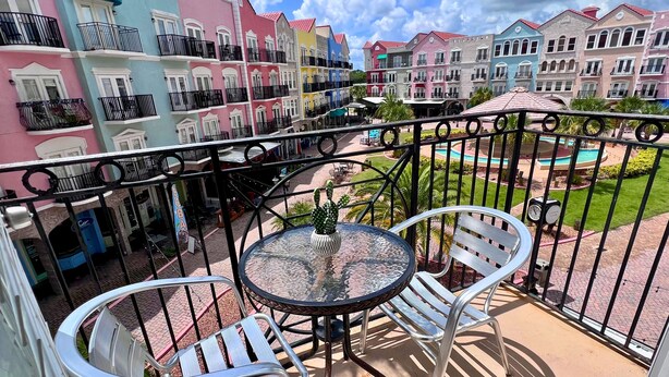 A balcony with two metal chairs and a glass table, overlooking colorful buildings and a pool area. - Elegant Condo - European Charm (Palm Coast)