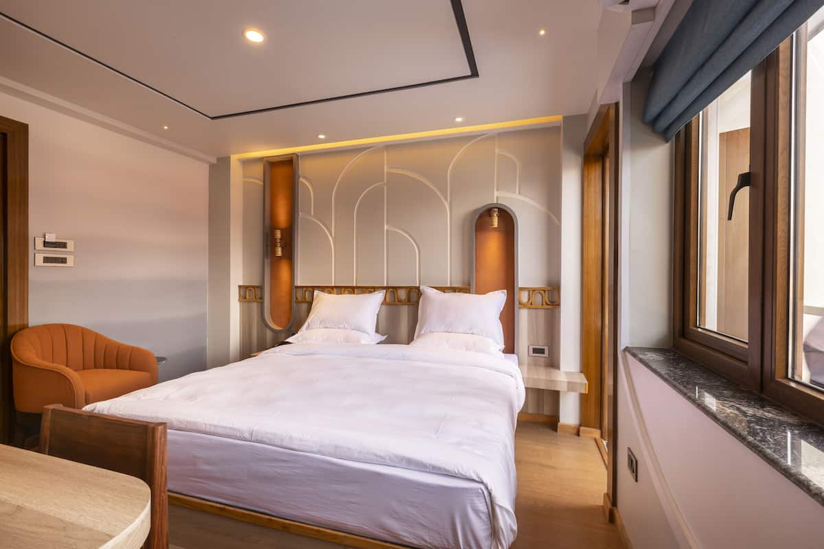 Ein modernes Hotelzimmer mit einem großen Bett, einem Holzstuhl, einem kleinen Tisch und einem Fenster mit Vorhängen.
