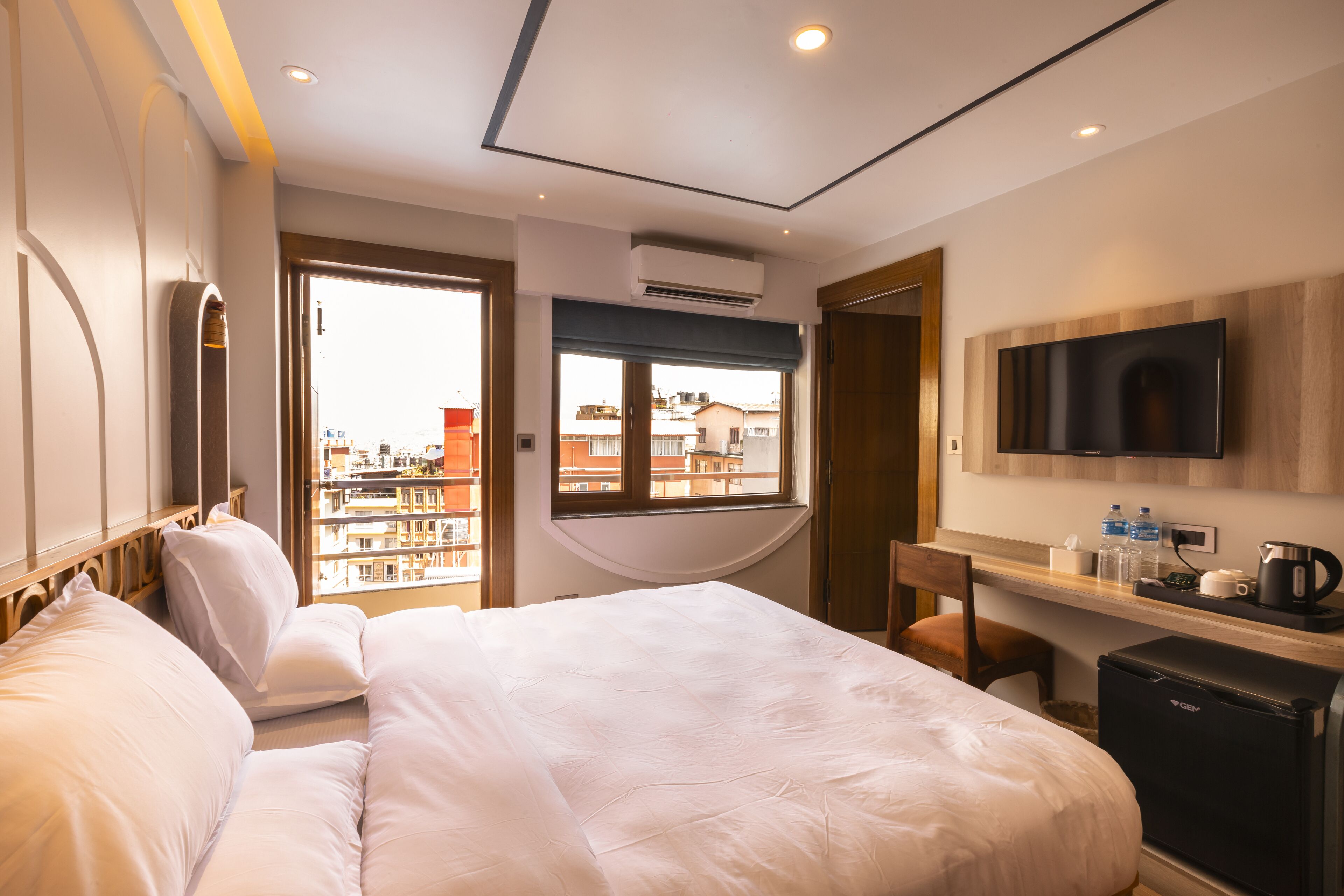 A hotel room with a large bed, a desk with a coffee maker, a TV, and a view of buildings through the window.