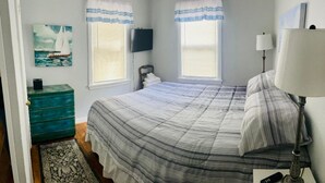 A bedroom with a large bed, two windows with curtains, a television on the wall, a lamp, and a painting on the wall.