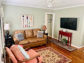 Una sala de estar con un sofá de cuero marrón, un sillón rosa, una mesita de noche roja, un televisor de pantalla plana y un ventilador de techo.