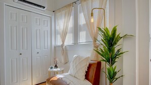 Une pièce avec un canapé en cuir marron, un plaid blanc, une plante en pot, une petite table ronde avec un vase, et une porte blanche surmontée d’une fenêtre.