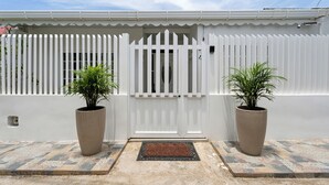 Une porte blanche ornée d’un motif décoratif, flanquée de deux grands pots de plantes, mène à une entrée carrelée.
