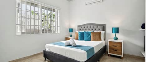 Un dormitorio con una cama, dos mesitas de noche, una ventana con rejas y un equipo de aire acondicionado.