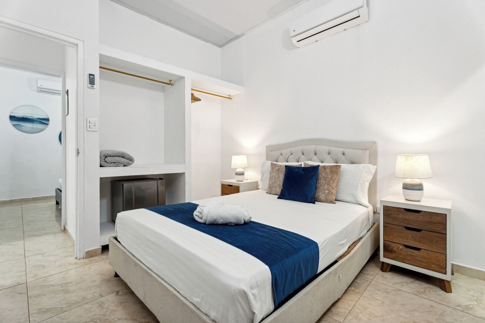 Quarto de hotel moderno com uma cama grande, mesas de cabeceira, ar condicionado e vista para o corredor.