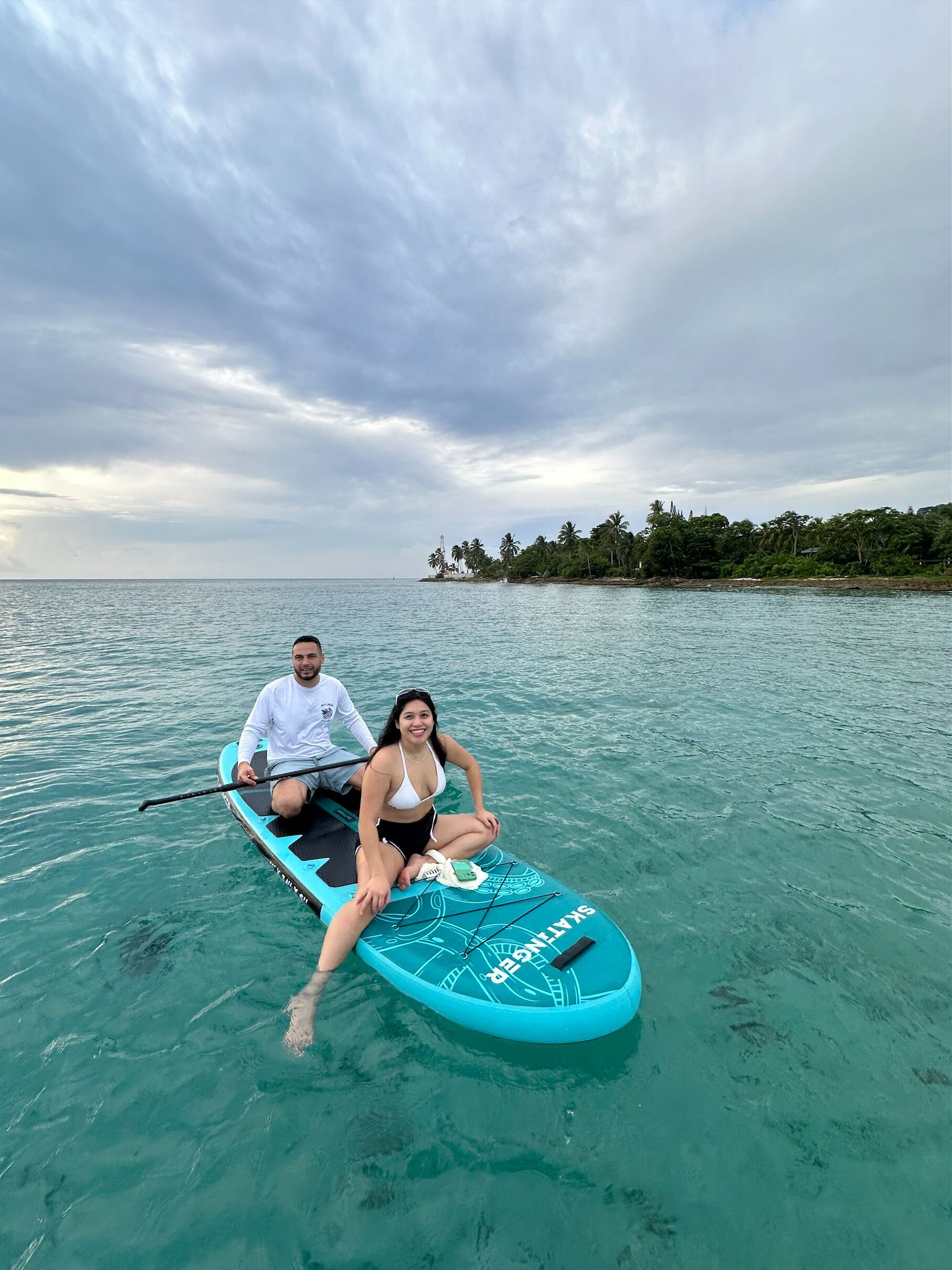 Two people on a paddleboard in clear water with a tropical island in the background.