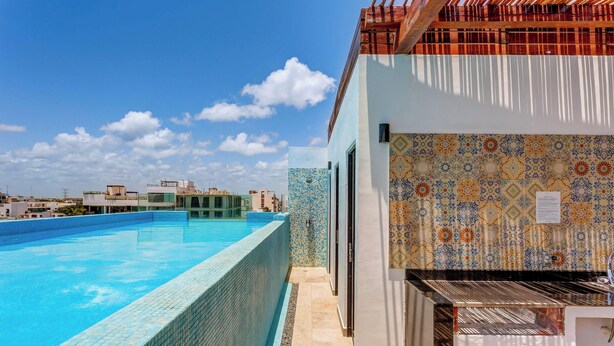 En swimmingpool med flisekant, en flisevæg med et dekorativt mønster og udsigt til omkringliggende bygninger.