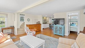 A spacious living room with a white coffee table, a wooden cabinet, and a television. There is a staircase leading to another level, and a door with a view of the outside.