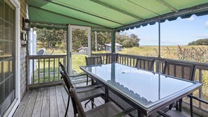 A covered outdoor area with a glass table and chairs, wooden deck, and a view of a grassy field and trees.