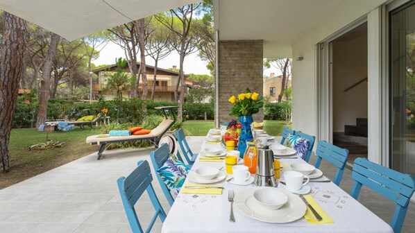 A dining area with a long table set for breakfast, outdoor seating, and a view of the surrounding garden and buildings. - La Villa in Pineta 10 Emma Villas (Castiglione della Pescaia)