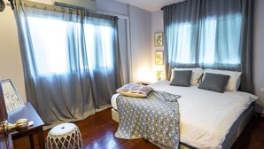 A bedroom with a large bed, grey curtains, a bedside table with a lamp, and a small stool.