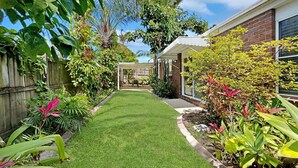 Un jardin bien entretenu, avec une pelouse, diverses plantes et une maison en briques dotée d’un patio couvert.
