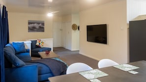 Una sala de estar con un sofá azul, una mesa de centro y una mesa de comedor con sillas. Hay un televisor montado en la pared y una puerta que da a otra habitación.