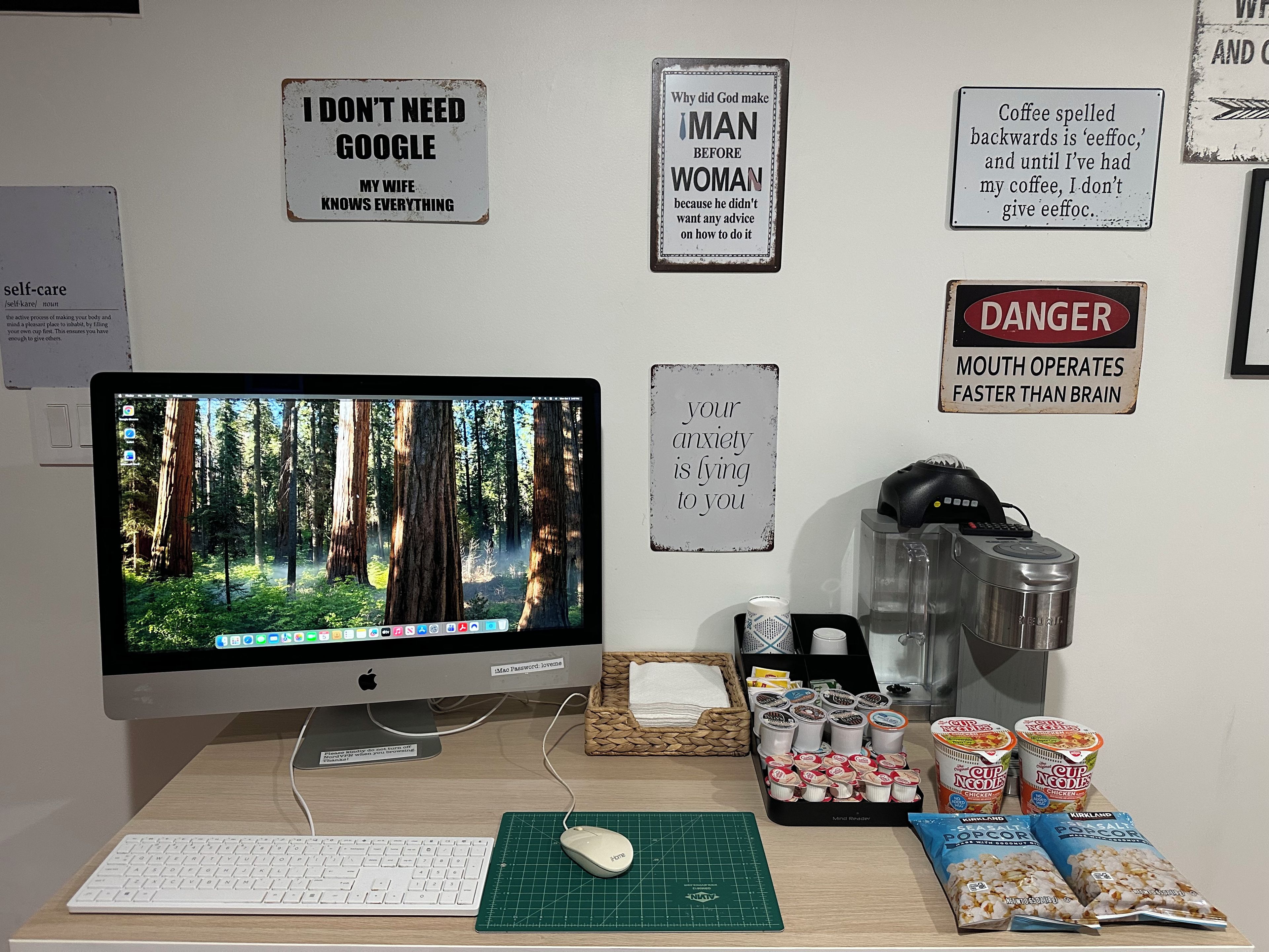A desk with a computer monitor, keyboard, mouse, and various items including a coffee maker, popcorn, and snack packages.