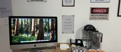 A desk with a computer monitor, keyboard, mouse, and various items including a coffee maker, popcorn, and snack packages.
