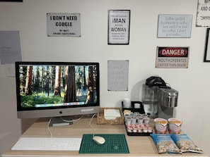 Un escritorio con un monitor de computadora, teclado, ratón y diversos objetos, incluyendo una cafetera, palomitas de maíz y paquetes de comida.