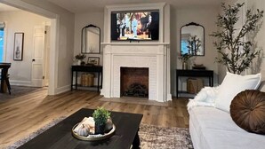 A living room with a fireplace, a ceiling fan, and a television. - Blue Moon Cottage: Historic Owensboro Home w/ Large Pool, Music Room, Location! (Owensboro)