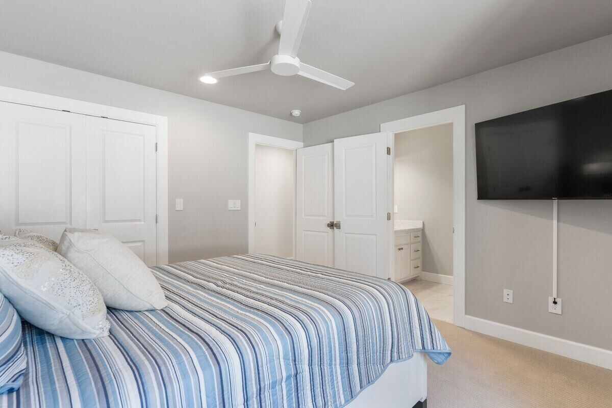 Un dormitorio con una cama, ventilador de techo, televisor montado en la pared y una puerta que da a otra habitación.