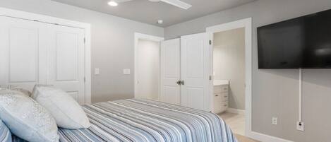 Un dormitorio con una cama, ventilador de techo, televisor montado en la pared y una puerta que da a otra habitación.