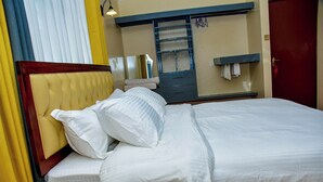 Un lit bien fait, avec du linge de lit blanc et une tête de lit en bois, une petite table de chevet et un miroir au-dessus d’une commode sur laquelle est accrochée une serviette.