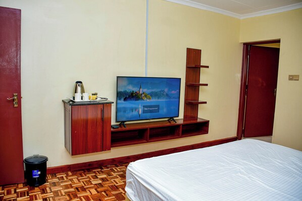 A hotel room with a bed, a television mounted on a wooden shelf, a coffee maker, and a trash bin.