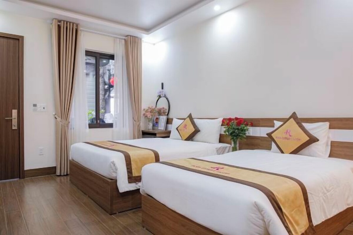 Ein Hotelzimmer mit zwei Betten, Holzboden, einem Fenster mit Vorhängen und einem Nachttisch mit Blumen in einer Vase.