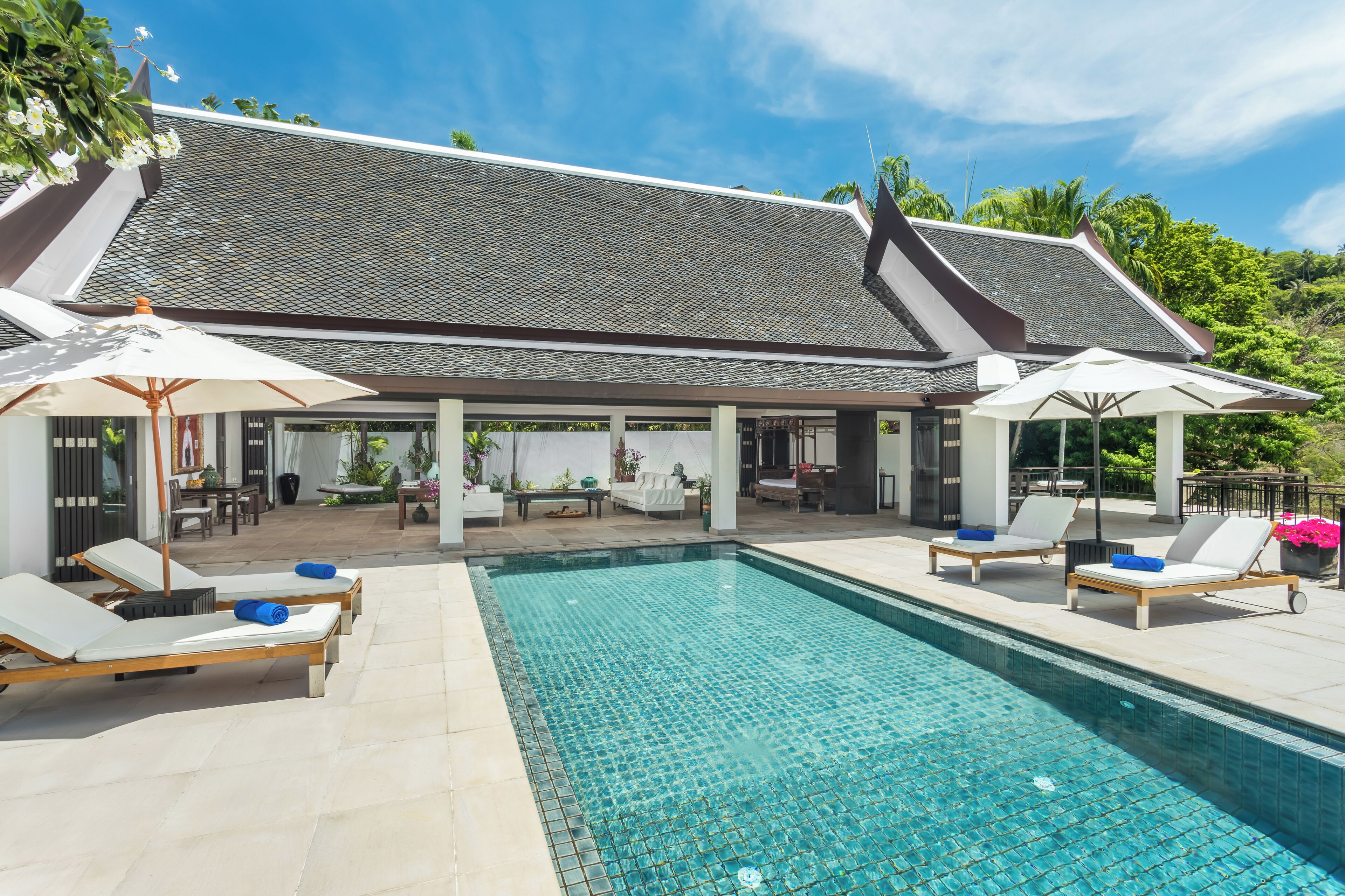 A poolside area with lounge chairs and umbrellas, a covered seating area, and a building with a tiled roof.