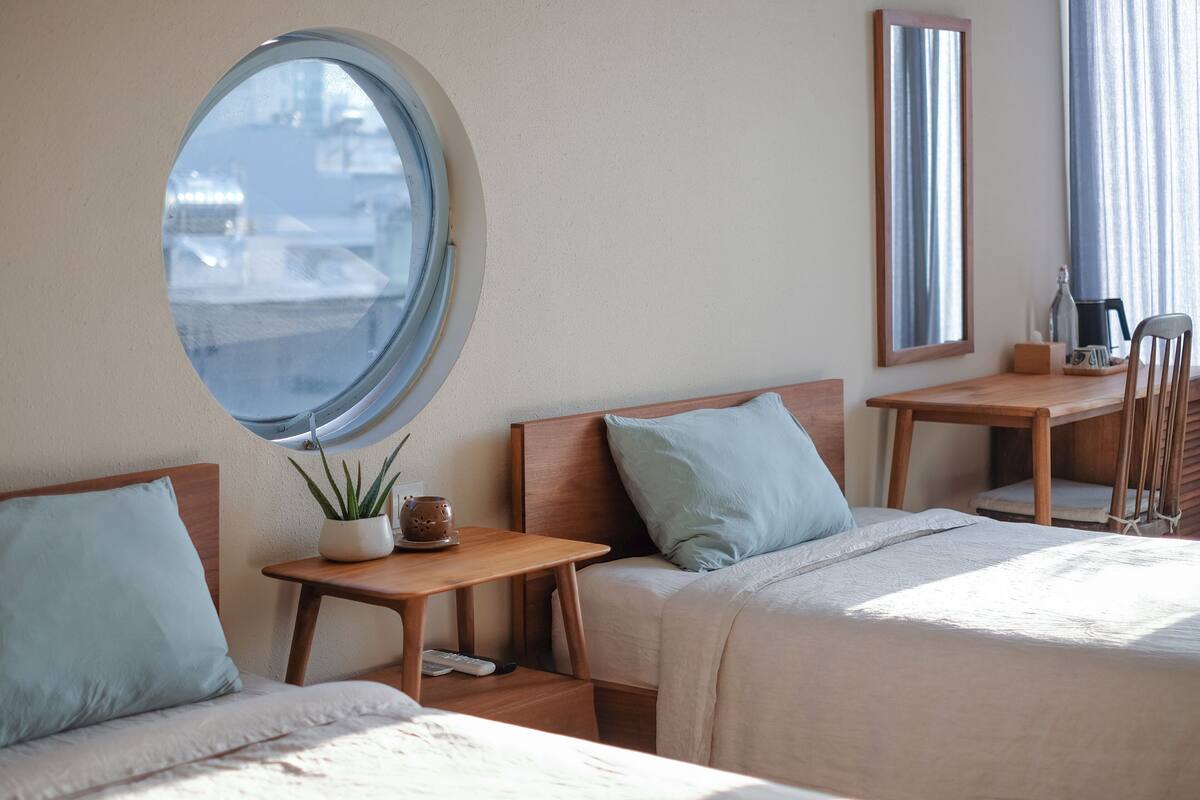 A hotel room with two beds, a round window, a small table with a plant, and a mirror on the wall.