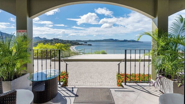 A covered outdoor area with a glass table and wicker chairs, overlooking a beach and ocean. - Luxury Condo with Panoramic Ocean Views, Sparkling Infinity Pool, and Modern Open-Concept Design (Playa Flamingo)