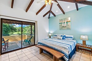 A bedroom with a large bed, a bench, a nightstand, a ceiling fan, and a view of greenery through a sliding door.