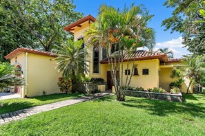 A two-story yellow building with a tiled roof, surrounded by lush greenery and palm trees.