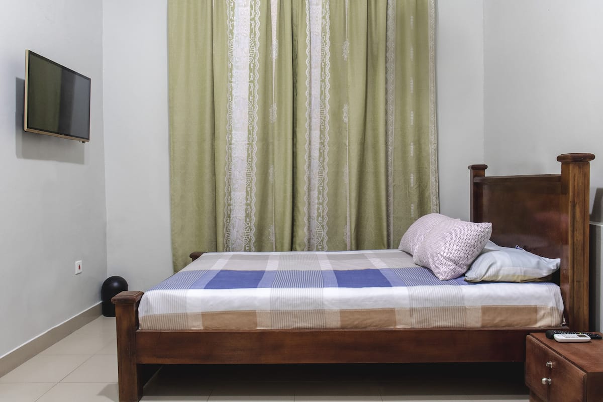 Un lit simple avec un cadre en bois, une couvre-lit à carreaux et deux oreillers. Un téléviseur est fixé au mur, sur la gauche.