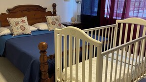 Quarto com cama de madeira, berço, mesa de cabeceira, termostato fixado na parede e janela com cortinas.