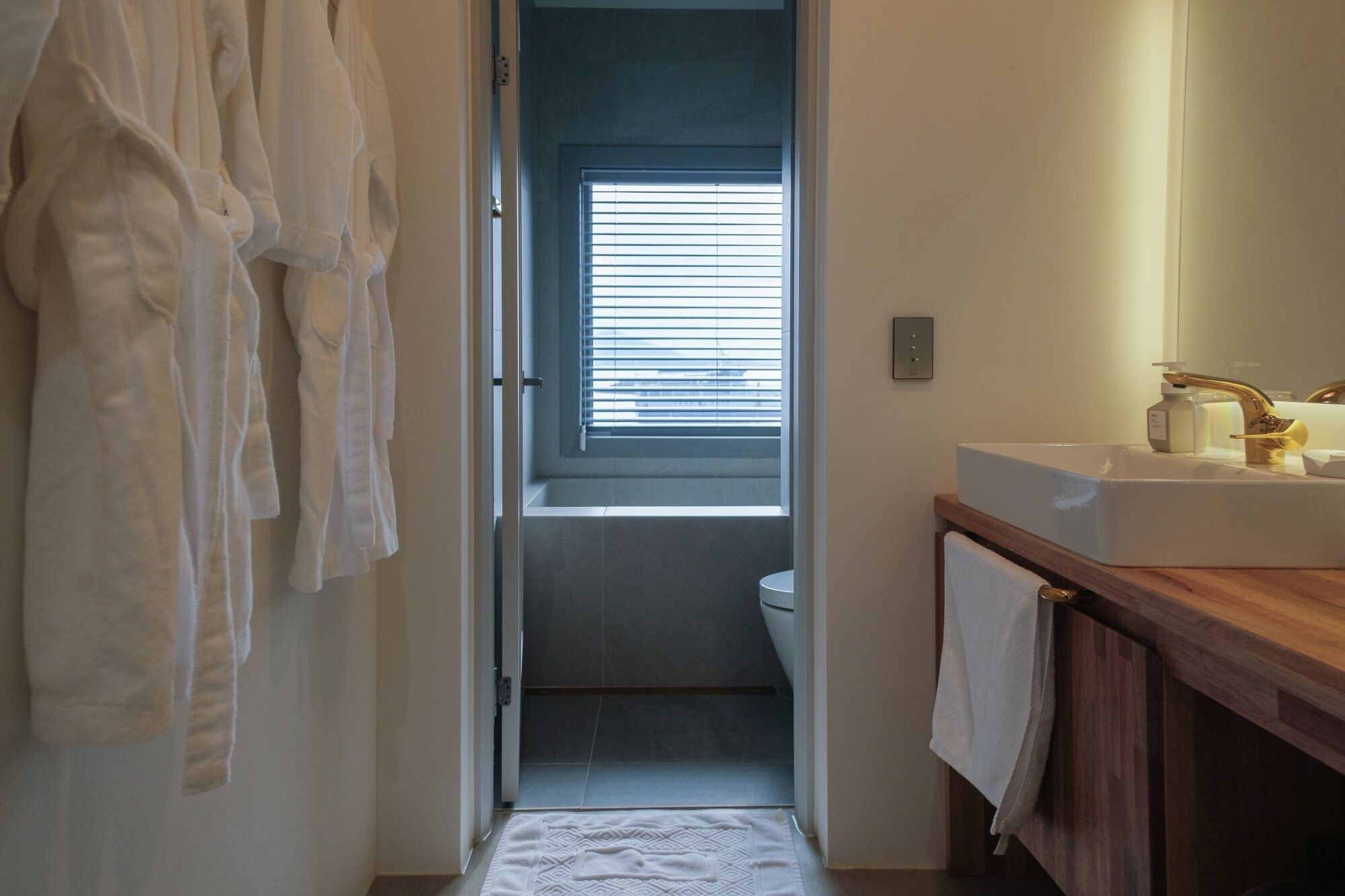 Une salle de bain avec un meuble-lavabo en bois, un lavabo blanc et une fenêtre avec des stores.