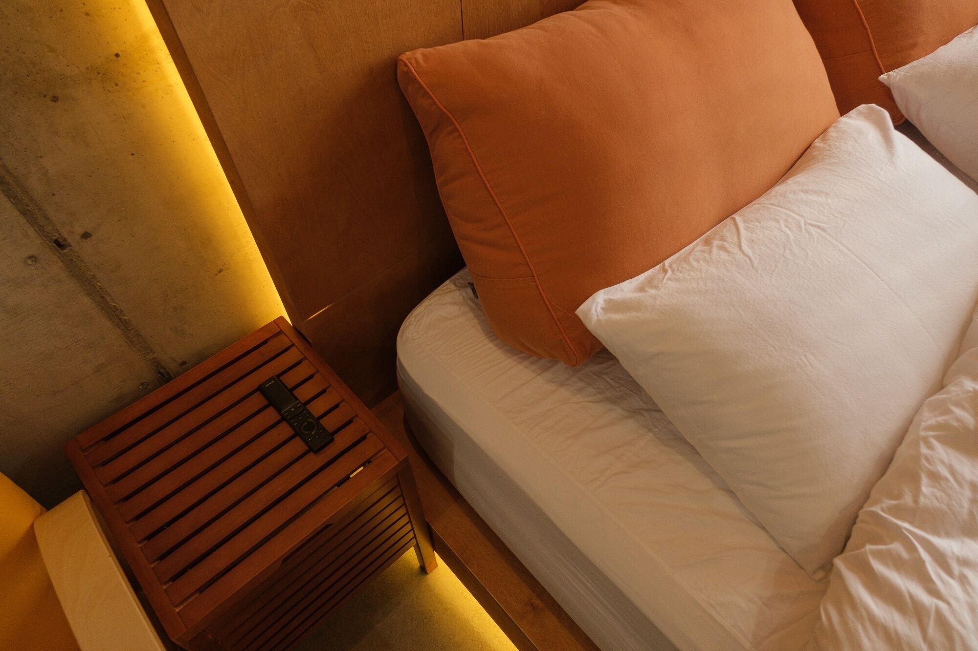 Un lit avec une literie blanche et un oreiller orange, une table de chevet en bois avec une télécommande, et un mur jaune.