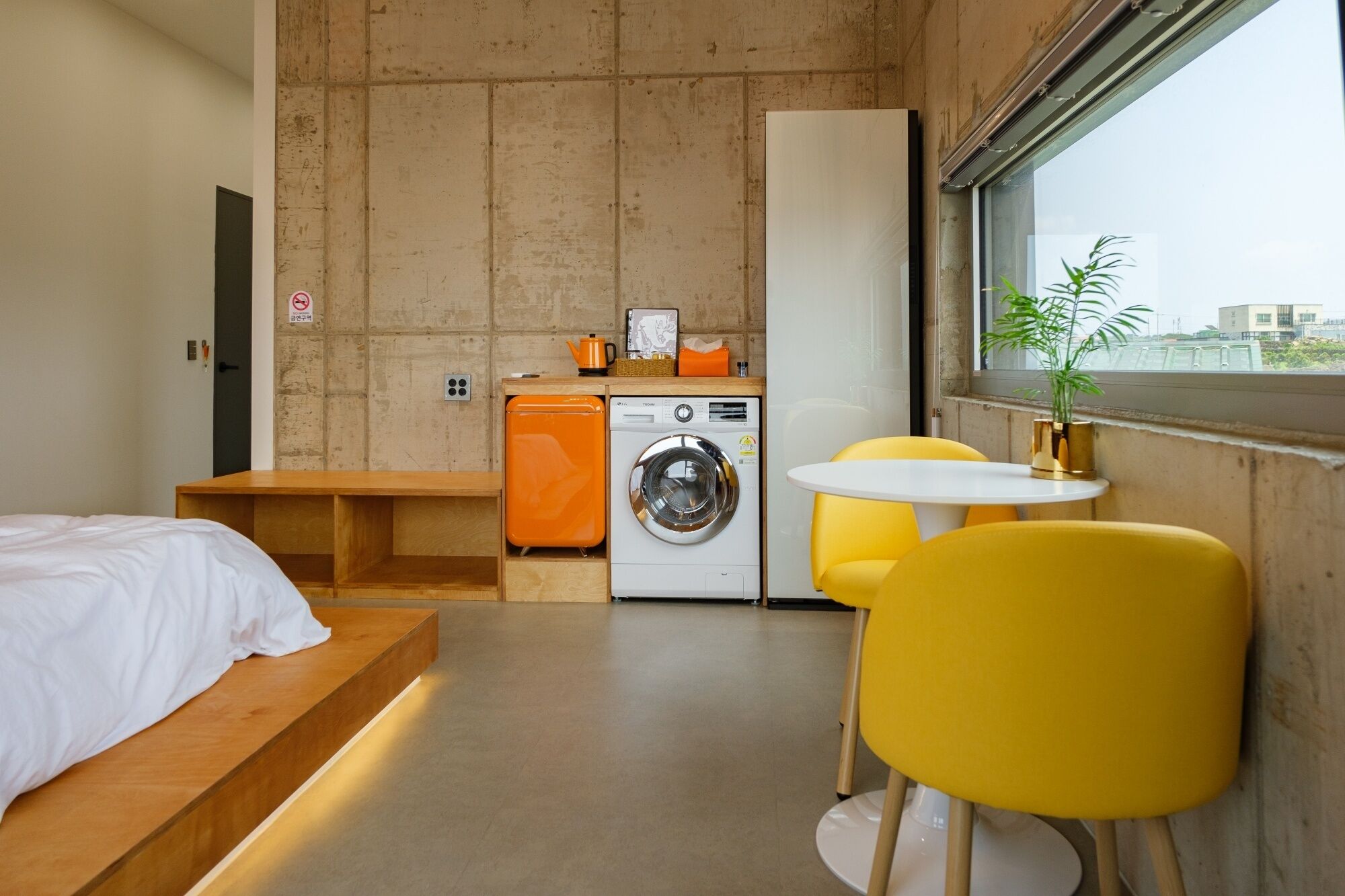A compact hotel room with a bed, a washing machine, a small table, and a chair.