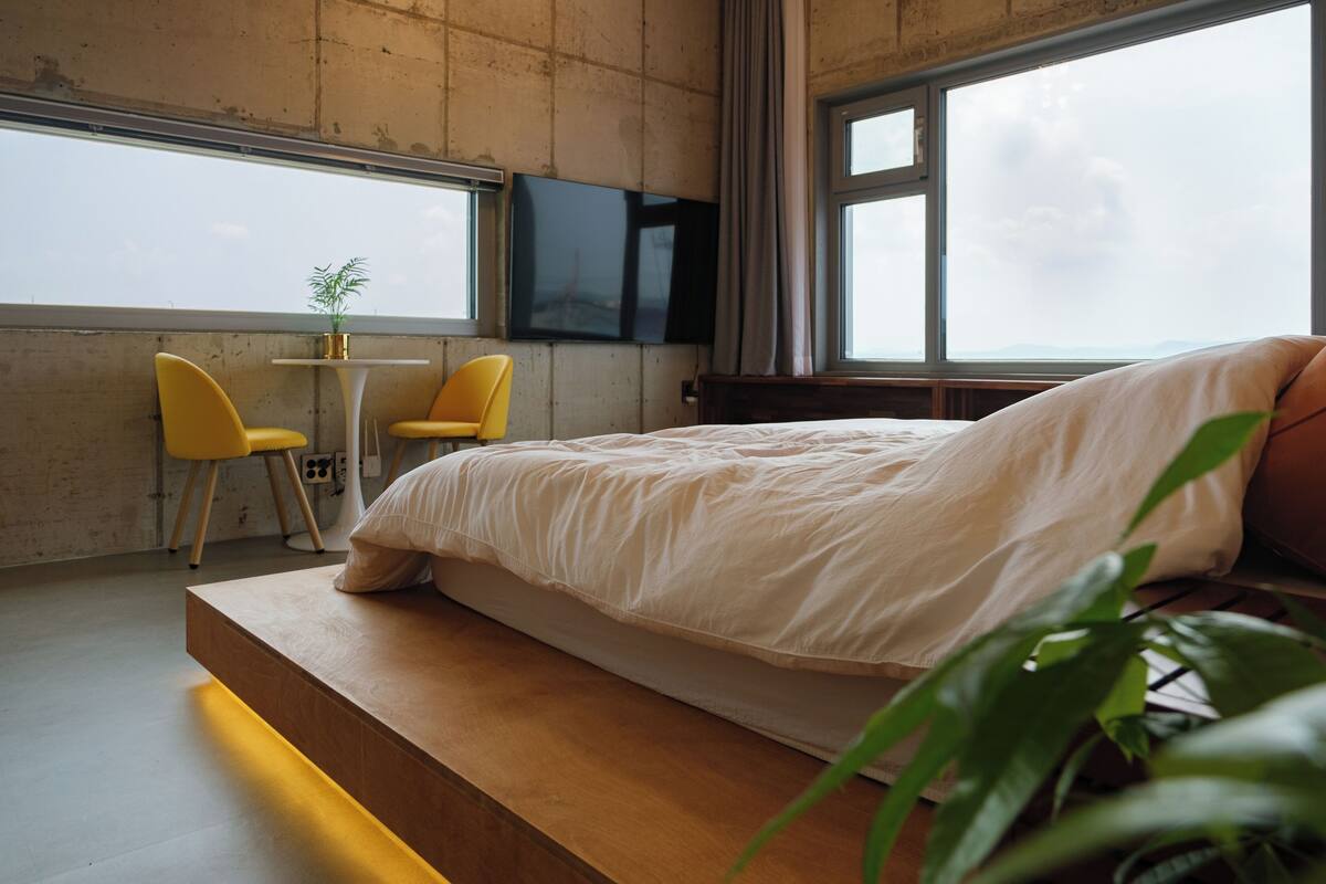 A modern bedroom with a large bed, a TV mounted on the wall, and a view of the sky through large windows.