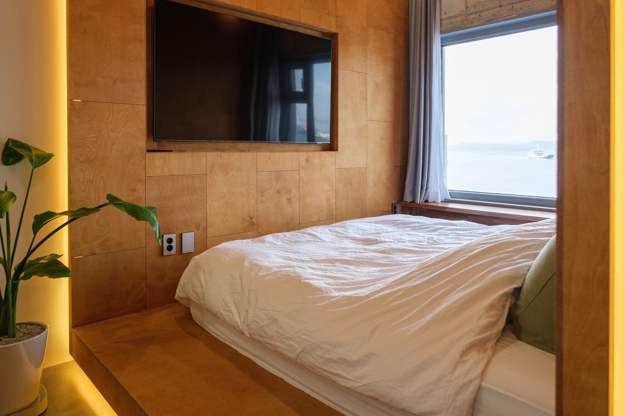 A bedroom with a bed, a TV mounted on the wall, a window with a view of the sea, and a potted plant.