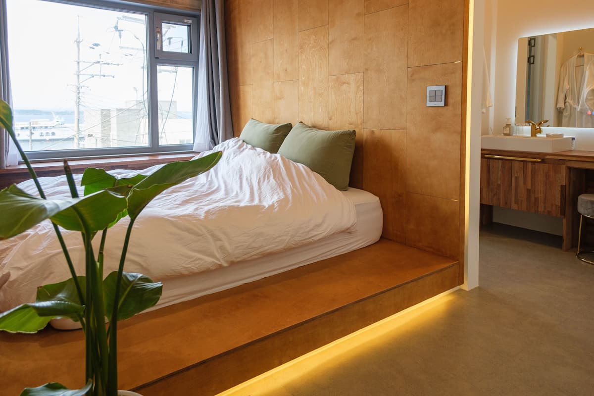 A modern bedroom with a wooden headboard, a large window, and a bedside plant.