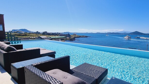 Una piscina con agua azul cristalina, rodeada de muebles de mimbre y con vistas a un cuerpo de agua con barcos.