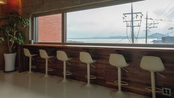 Un área de bar moderna con una barra de madera y cinco taburetes blancos. Un ventanal ofrece vistas a un paisaje costero con líneas eléctricas y una cadena montañosa distante.