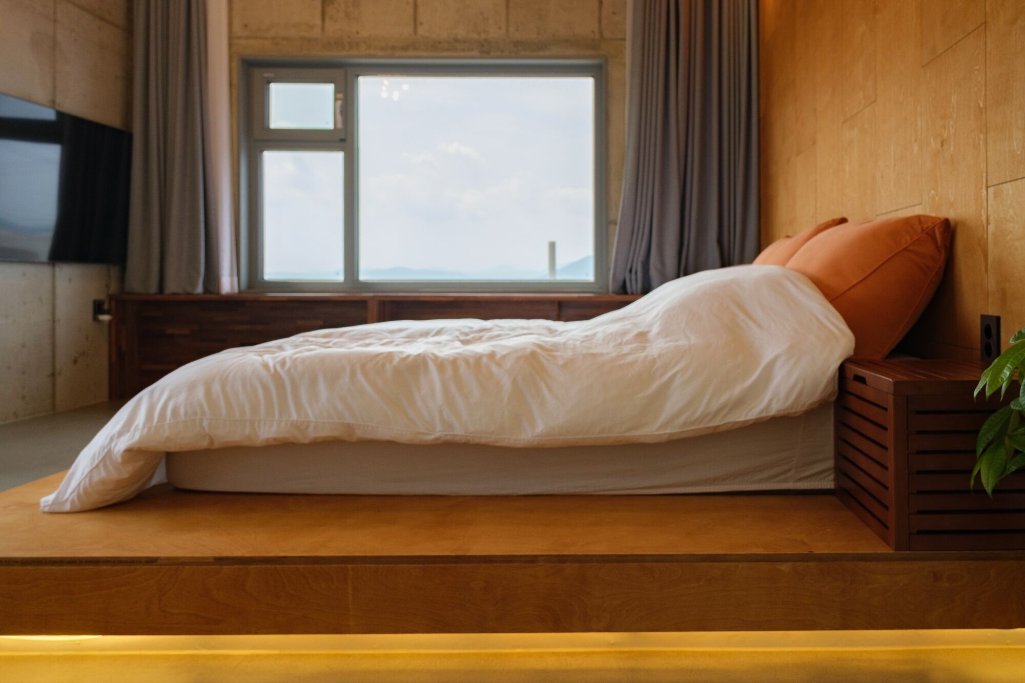 A bed with a white mattress and orange pillow, positioned against a wooden wall with a window above.