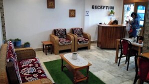 Un área de recepción con un mostrador, dos sofás de mimbre con cojines estampados, una pequeña mesa con un mantel blanco y una alfombra de césped artificial verde.
