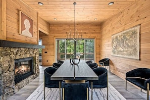 Un intérieur moderne agrémenté d’une cheminée en pierre, d’une longue table à manger avec des chaises, et de grandes fenêtres offrant une vue sur l’extérieur.