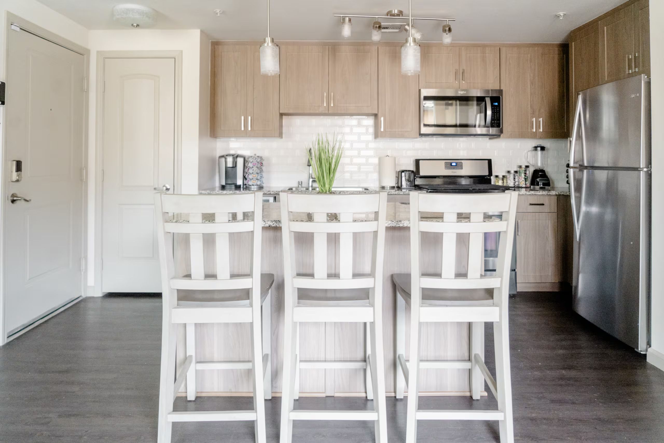A modern kitchen with three white bar stools, a stainless steel refrigerator, a microwave, and a coffee maker.