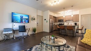 A modern living room with a glass coffee table, a flat-screen TV mounted on the wall, a kitchen area with wooden cabinets and stainless steel appliances, and a seating area with a brown leather sofa.