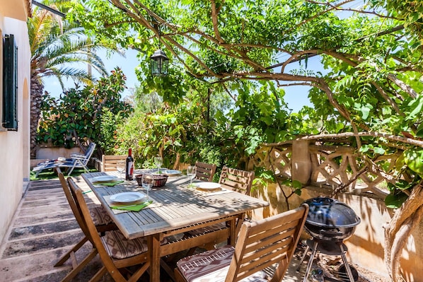 An outdoor dining area with a wooden table and chairs, a grill, and a view of lush greenery.